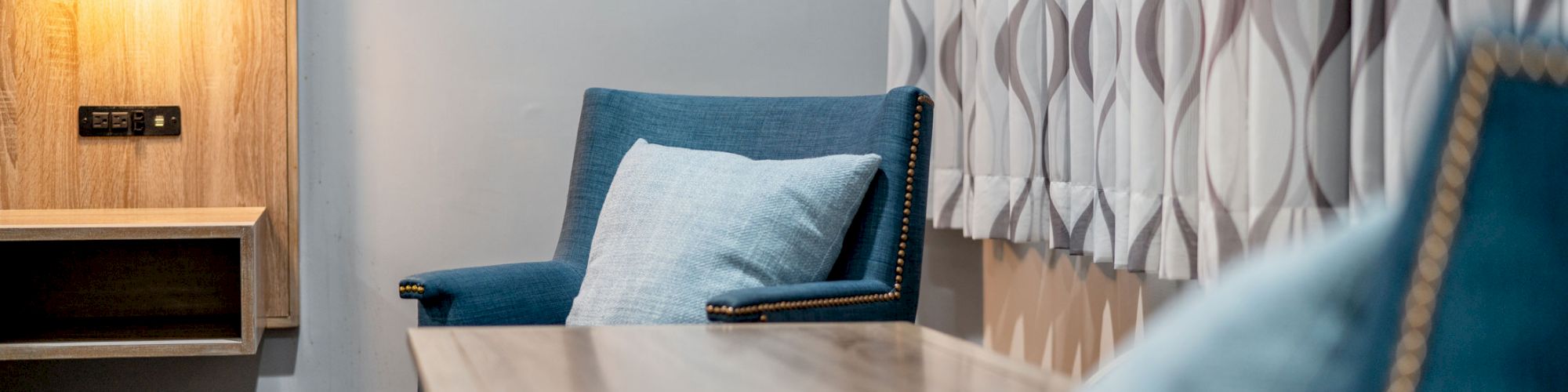 The image shows a room with a wooden table, two blue chairs with light blue pillows, a lit wall sconce, and patterned curtains.