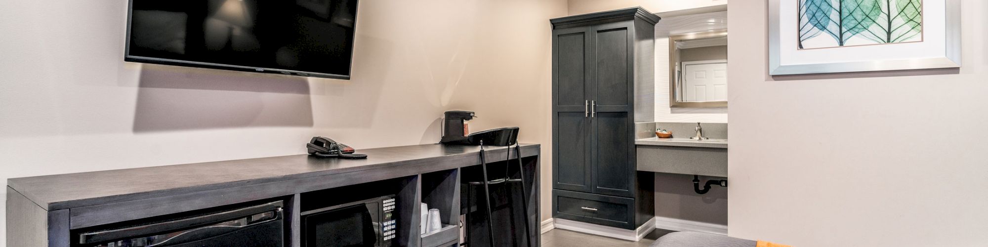 A modern hotel room with a wall-mounted TV, desk, chair, mini-fridge, and closet. The room features neutral colors and a simple design.