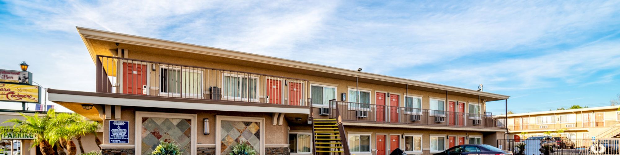 The image features a two-story motel with exterior staircases, parked cars, and a clear blue sky in the background.