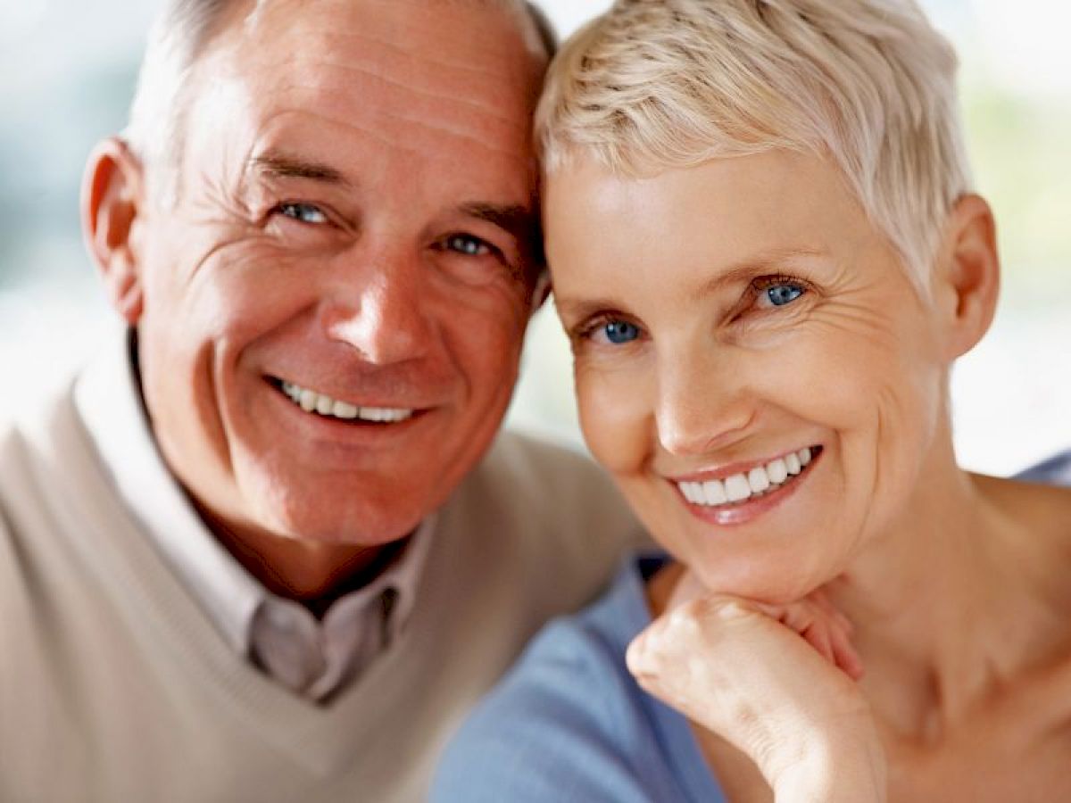 An elderly couple is smiling warmly at the camera, closely leaning towards each other, capturing a moment of happiness and togetherness.