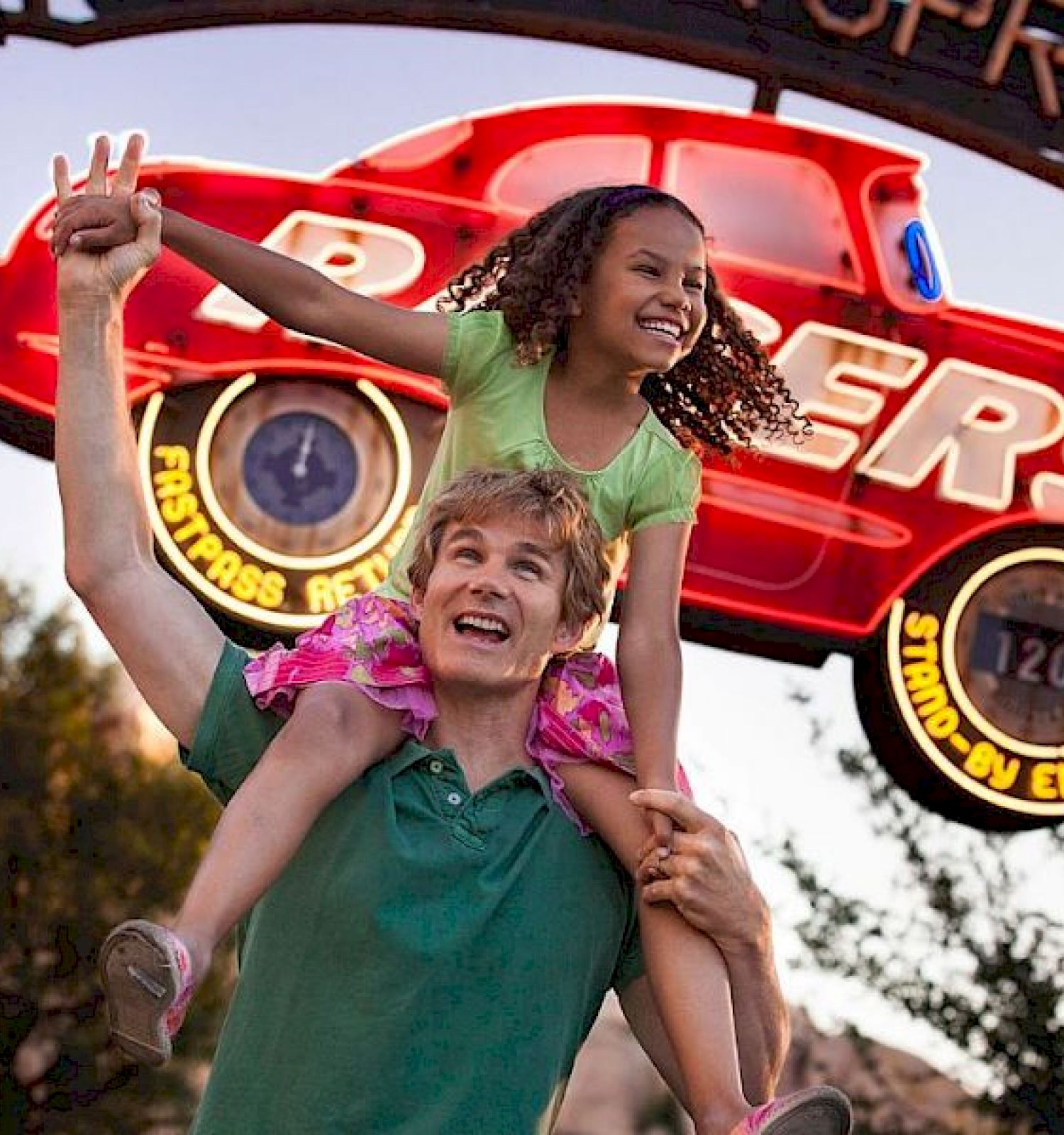 A man carries a smiling girl on his shoulders in front of a 'Cars' themed attraction, featuring a large sign with a red car and cheerful atmosphere.