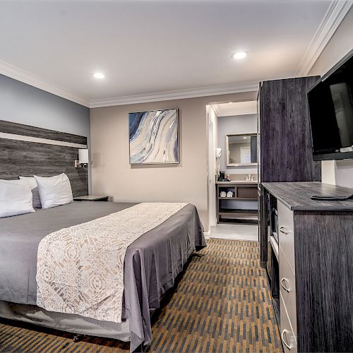 This image shows a neatly arranged hotel room with a bed, TV, and dresser, leading to a bathroom.