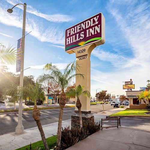 A street view showing the sign for "Friendly Hills Inn" beside the road, with palm trees, a few buildings, and a clear blue sky in the background.