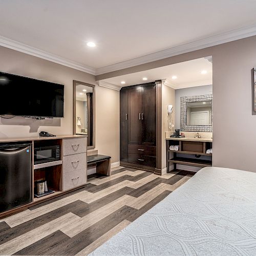 This image shows a modern hotel room with a TV, mini-fridge, microwave, dresser, and a bed. There is also a closet and bathroom vanity.