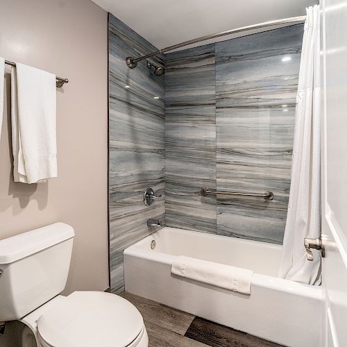 A modern bathroom with a toilet, a bathtub with gray-tiled walls, a shower curtain, and towels hanging on a rack.