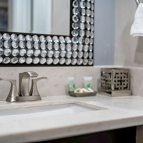 A bathroom sink area with a modern faucet, a decorative mirror, toiletries, and a towel holder with a white towel.