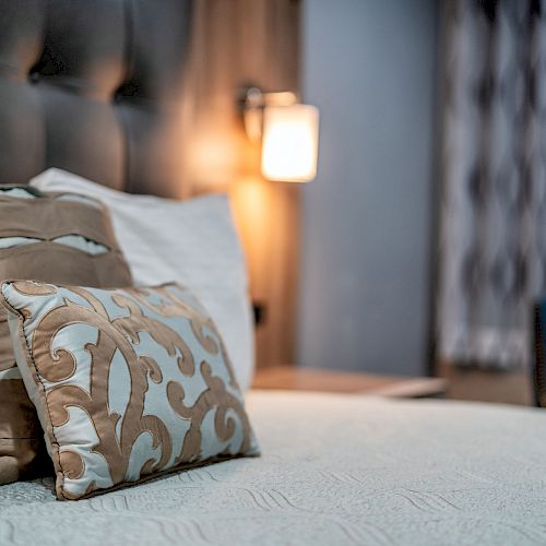 A neatly made bed with decorative pillows, a tufted headboard, and a lit wall lamp in a warmly lit room with patterned curtains.