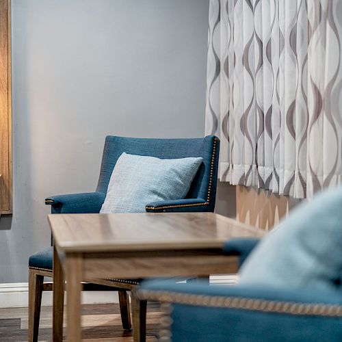 A cozy room with two blue chairs, a wooden table, a wall lamp, and patterned curtains next to a gray wall.