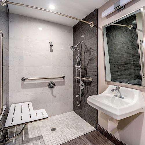 The image shows an accessible bathroom with a shower area, foldable shower chair, grab bars, a sink, and a mirror.