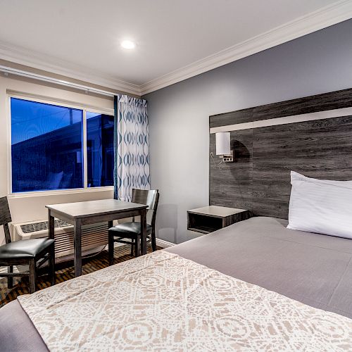 This image shows a modern hotel room with a bed, two chairs, a small table, a window with curtains, and a nightstand.