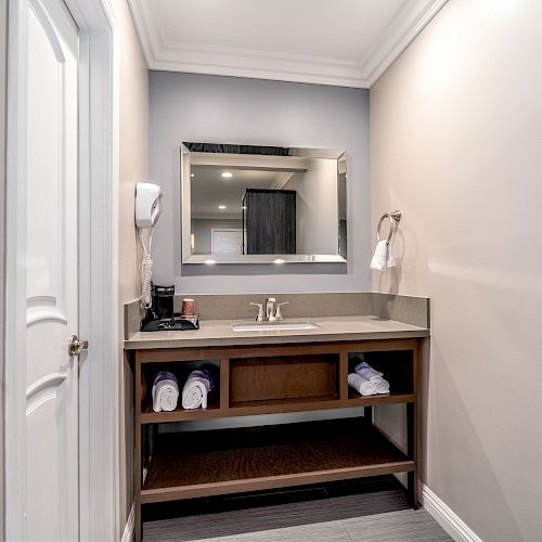 A bathroom vanity with a mirror, towels, hairdryer, and toiletries, set against light-colored walls with crown molding and a door to the side.