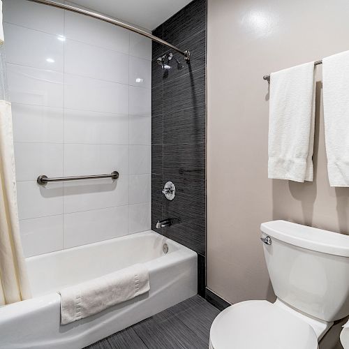 The image shows a modern bathroom with a bathtub and shower combo, shower curtain, grab bar, toilet, and towels hanging on a rack above the toilet.