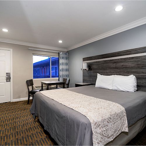 A modern hotel room with a large bed, white bedding, nightstands, a window, a small table with chairs, and recessed ceiling lights.