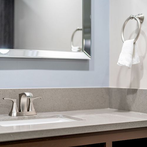 A modern bathroom sink with a faucet, a gray countertop, a wall-mounted mirror, and a towel ring with a white towel.