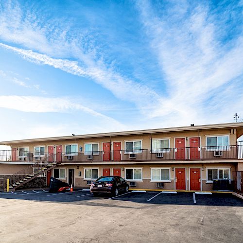 A two-story motel with red doors, parked cars, and an empty parking lot under a blue, partly cloudy sky.