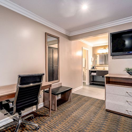 The image shows a hotel room with a desk, chair, mirror, TV, dresser, plant, and an open door to a bathroom.