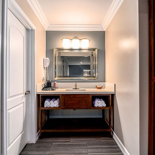 A bathroom with a sink, mirror, three-light fixture, towels on a shelf below the sink, a door to the left, and wooden cabinets to the right.