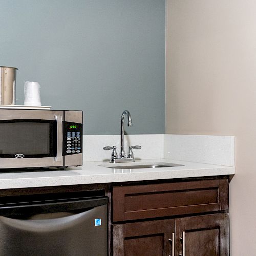 The image shows a small kitchenette with a microwave, mini-fridge, sink, and brown cabinets, featuring a metallic ice bucket and disposable cups on the counter.