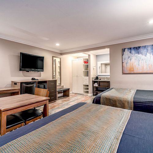 The image shows a modern hotel room with two beds, a wall-mounted TV, a table with chairs, a dresser, and a visible bathroom area in the background.