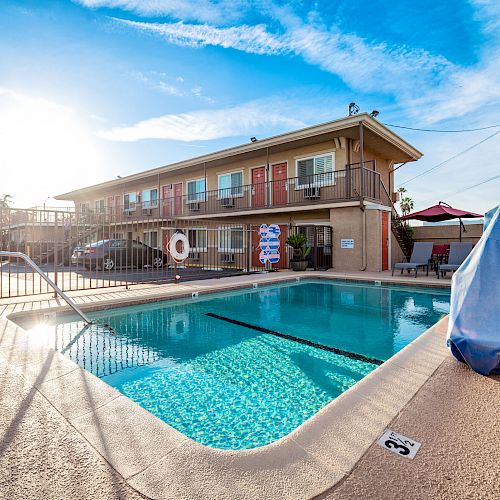 An outdoor pool area with ladders, clear water, and a fenced boundary is in front of a two-story building under a blue sky with scattered clouds.