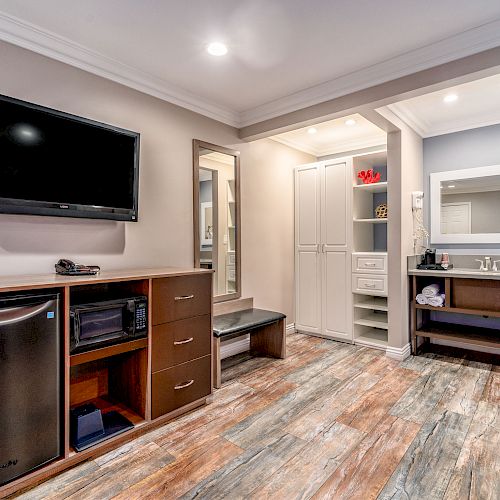 The image shows a modern hotel room with a mounted TV, wooden floors, a mini-fridge, storage units, and a bathroom vanity area with a large mirror.