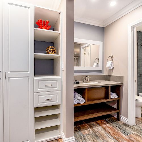 This image shows a modern bathroom with storage cabinets, a vanity with a large mirror, and an adjacent area featuring a toilet and shower.