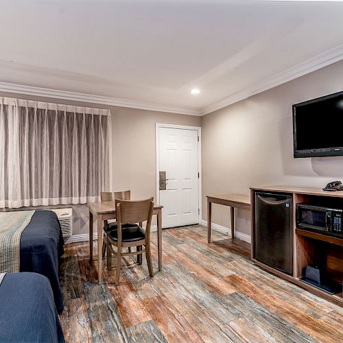 A hotel room with two beds, a small dining table with chairs, a wall-mounted TV, and a cabinet with a fridge and microwave.