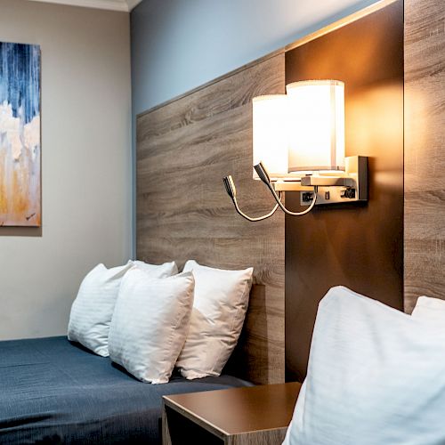 A modern hotel room with a bed, white pillows, wood-paneled headboard, wall-mounted lamp, and abstract artwork in the background.