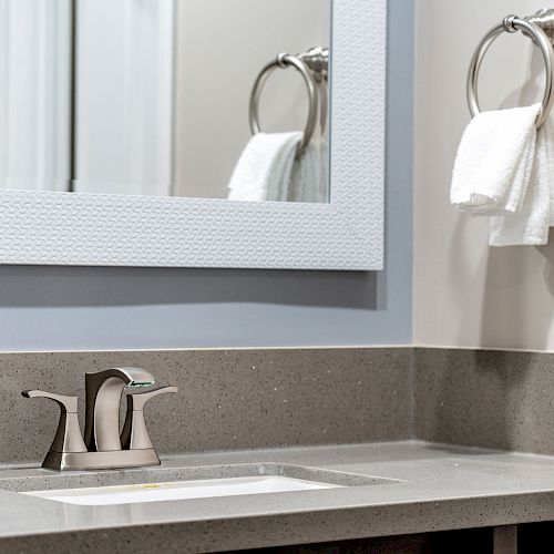 The image shows a bathroom sink with a modern faucet, a large framed mirror, and a hand towel hanging on a ring next to the mirror.