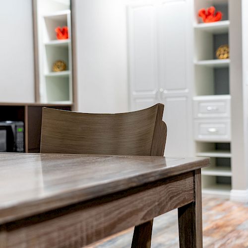 The image shows a modern room with a wooden table, chair, wall shelves, a TV, and a kitchenette with a microwave, storage cabinets, and decorative items.