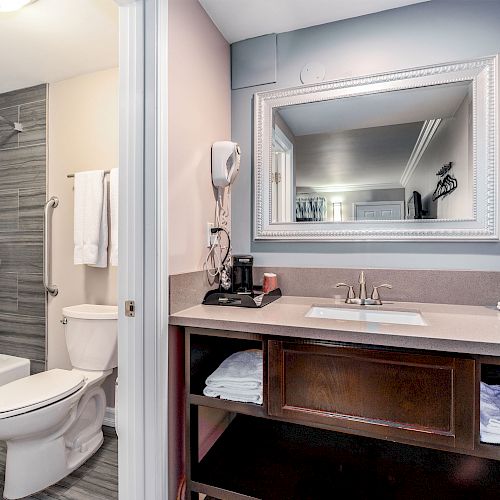 The image shows a clean bathroom with a bathtub, toilet, and a sink area with a mirror, towels, and toiletries.