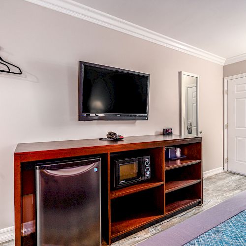 A hotel room featuring a TV on the wall, a dresser with a mini-fridge, microwave, and a mirror near the entrance door.