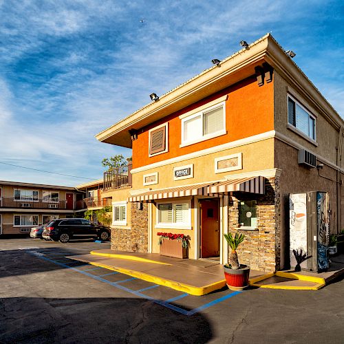 The image shows a two-story motel building with parking spaces and cars on a bright day, with a clear blue sky in the background.