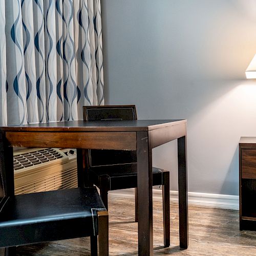 The image shows a small dining area with a dark wooden table, two chairs, an air conditioner, an end table, and a lamp beside patterned curtains.