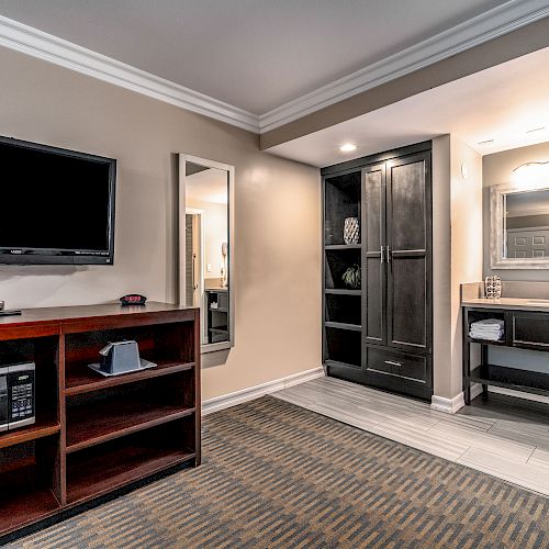 The image shows a modern hotel room with a TV, a mirror, a storage unit, and a small kitchenette area with a microwave and mini-fridge.