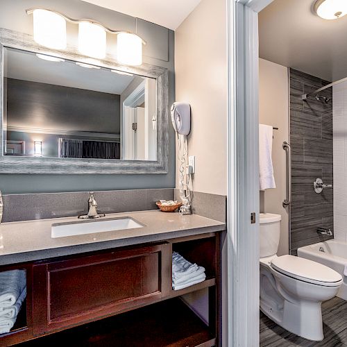 This image shows a modern bathroom with a sink, mirror, toilet, and bathtub, with towels neatly stored under the sink.