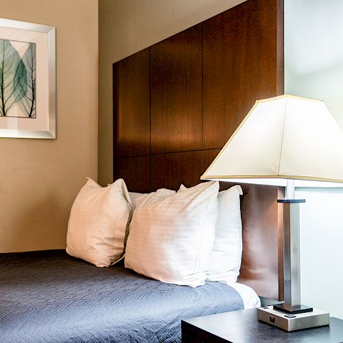 The image shows a neatly made bed with three white pillows, a bedside table, a modern lamp, and a picture of leaves on the wall.