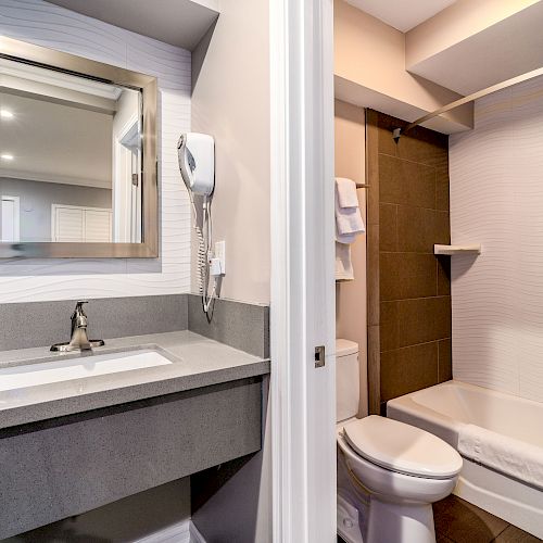 The image shows a modern bathroom with a gray vanity, large mirror, wall-mounted hairdryer, and a shower-tub combination with a white and brown tiling.