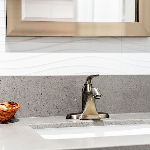 A modern bathroom sink with a metallic faucet, a rectangular mirror above, and a basket containing small bottles placed on the left side of the counter.