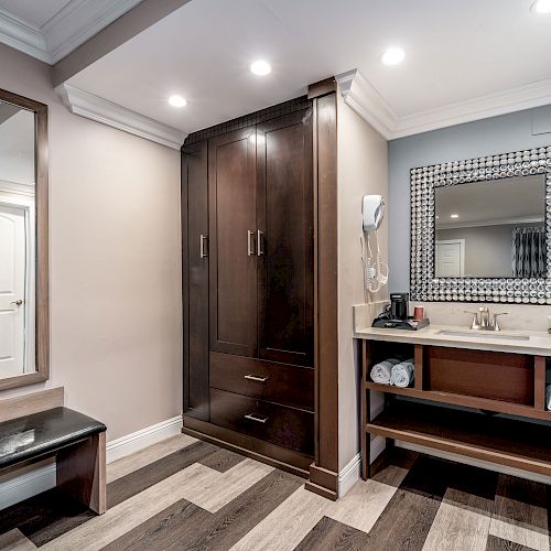 A stylish room features a large mirror, bench, wardrobe, and vanity with a coffee maker, neatly stacked towels, and a wall-mounted hairdryer, ending the sentence.