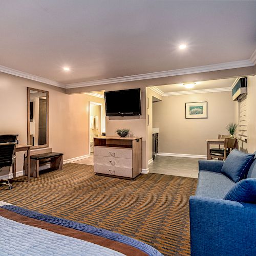 A hotel room with a sofa, bed, work desk, and TV. There is a dining area in the background and neutral decor.