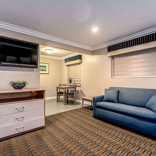 The image shows a hotel room with a sofa, a TV mounted on the wall, a chest of drawers, a dining table, and window blinds.