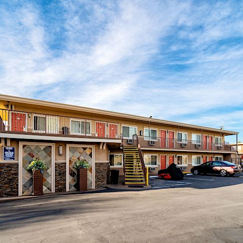 This image shows a two-story motel with exterior corridors, a central staircase, and a few parked cars, under a partly cloudy blue sky.