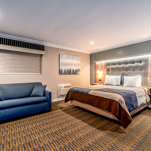 A comfortable hotel room features a large bed, blue sofa, modern décor, stylish lighting, and a wall-mounted air conditioner.