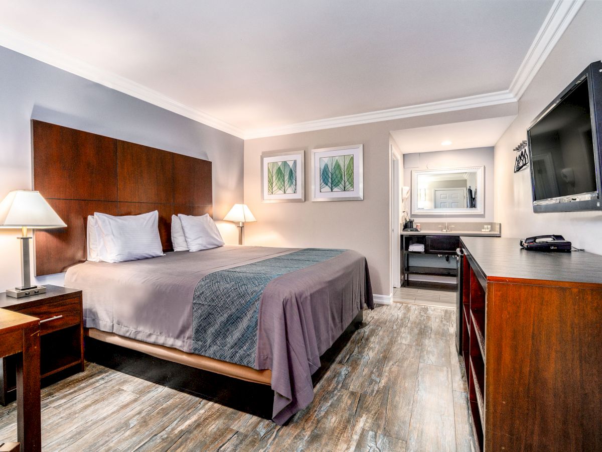 A modern hotel room with a king-size bed, two nightstands with lamps, a TV, wooden dresser, and a desk with a mirror in an alcove.