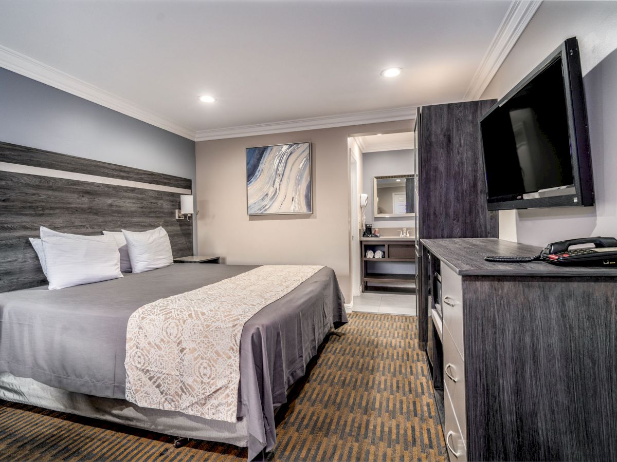 A modern hotel room with a king-size bed, a TV, a dresser, and a painting on the wall. The bathroom entrance is visible in the background.