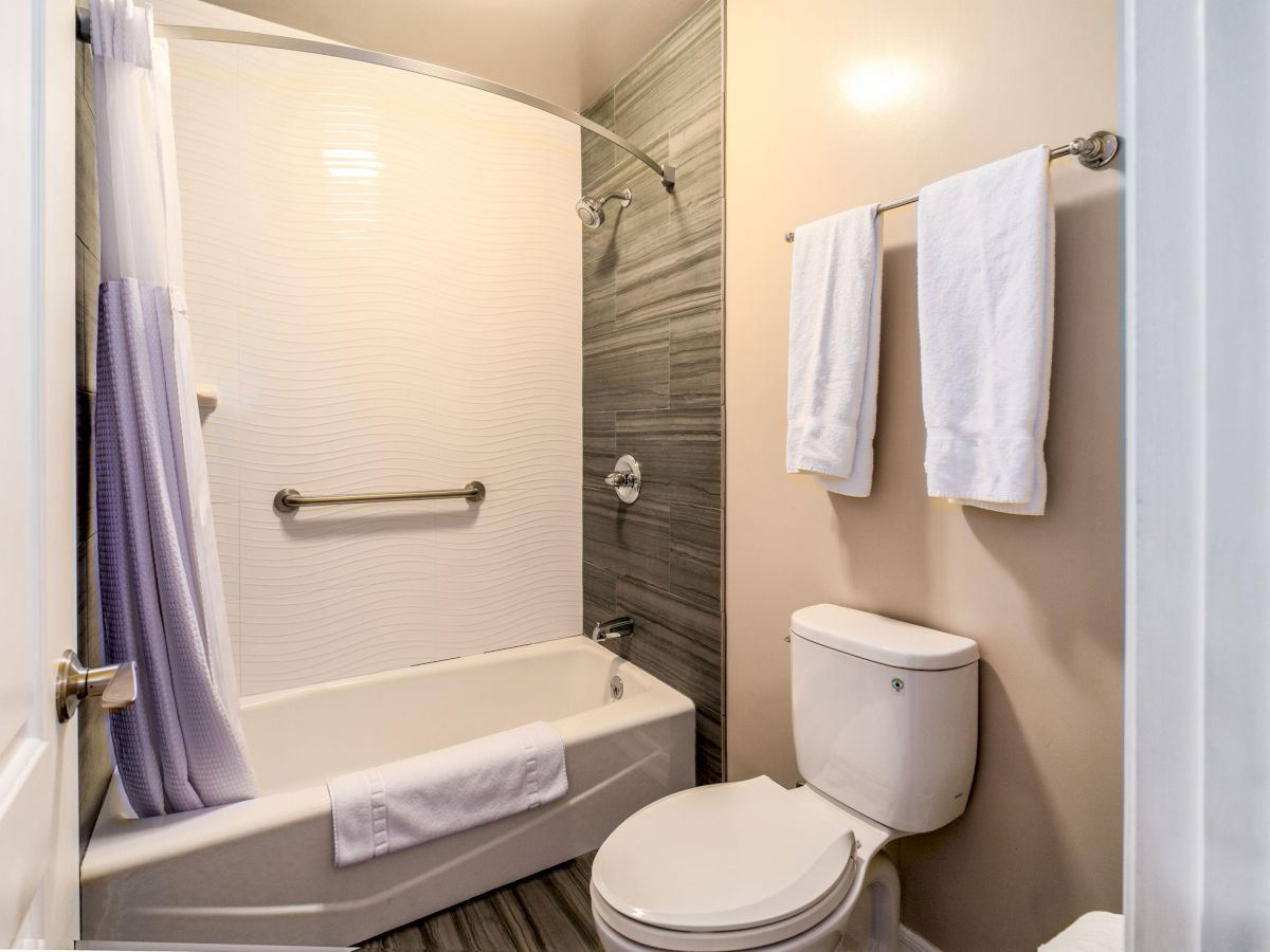 A small, neat bathroom featuring a bathtub with a shower, a toilet, a towel rack with white towels, and a modern design on the wall tiles.