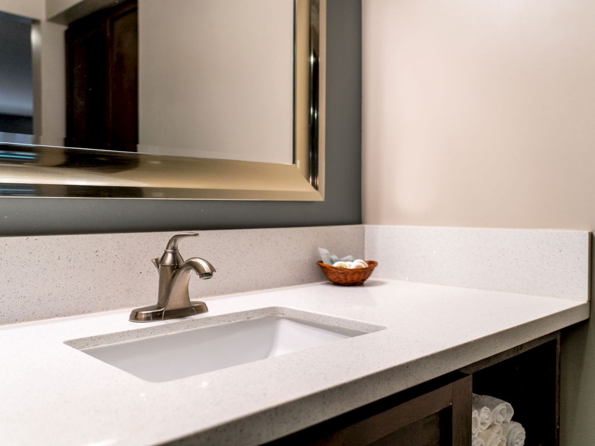 A modern bathroom sink with a white countertop, a polished metal faucet, a basket of toiletries, and a large mirror above the sink.