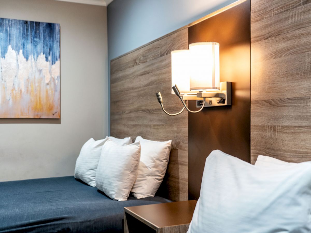 A modern bedroom with a bed, white pillows, a wooden headboard, a wall-mounted lamp, and a colorful painting on the wall.