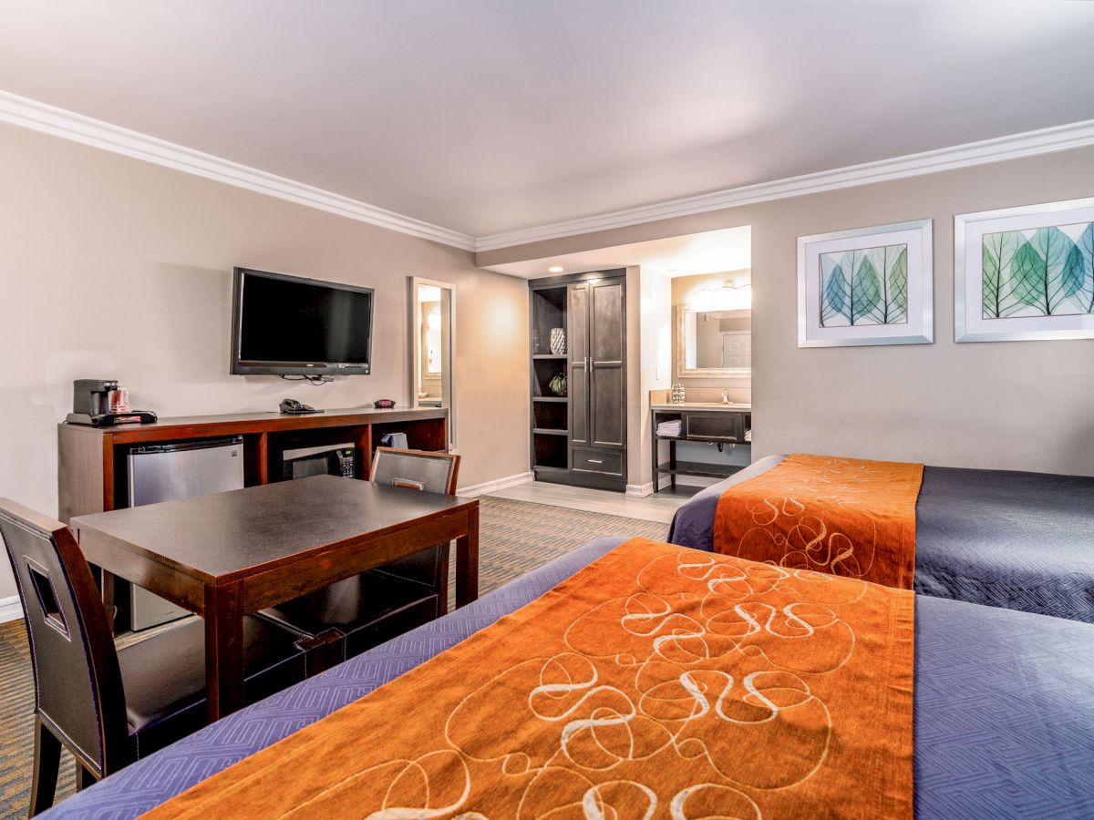 The image shows a hotel room with two beds, a TV, a small dining table with two chairs, framed art on the wall, and a bathroom area in the background.